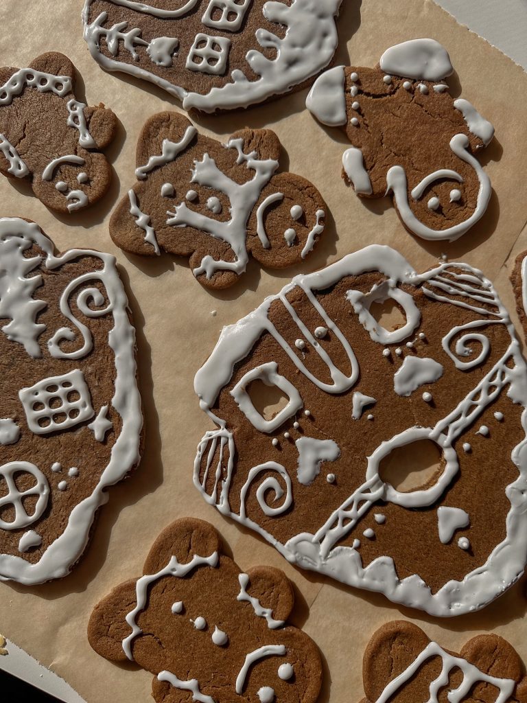 Gingerbread houses and gingerbread men spread across a kitchen table with bowls of frosting, sprinkles, and candy as young children decorate together during a Christmas baking tradition.